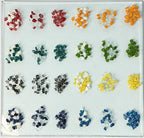 Assorted small colored glass gemstones arranged in a grid on a sheet of glass sitting on a light gray table top