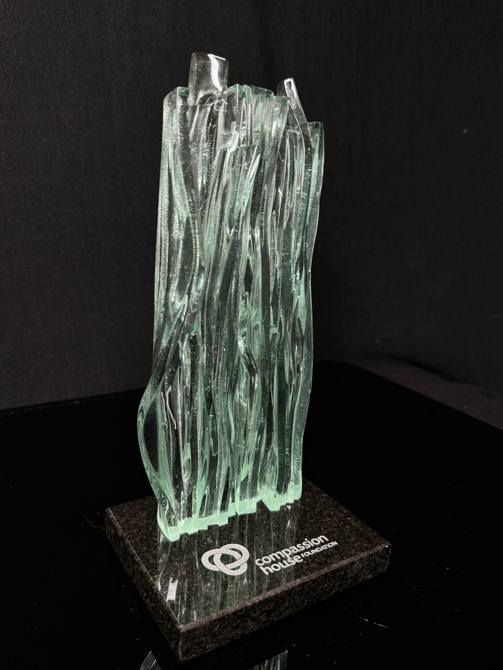 10 inch tall Canadian handcrafted Kiln Formed casted Glass waterfall award statue, that sits on top of a granite natural stone base with silver etching on top. the award stands in front of a black studio background