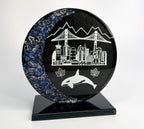 6 inch tall handcrafted glass award that has colored glass gems fused into the front of the award. the award is painted metallic black on the reverse with etching of Vancouver landmarks, Canadian maple leafs and an orca killer whale. the award stands on a black glass base in front of a studio background