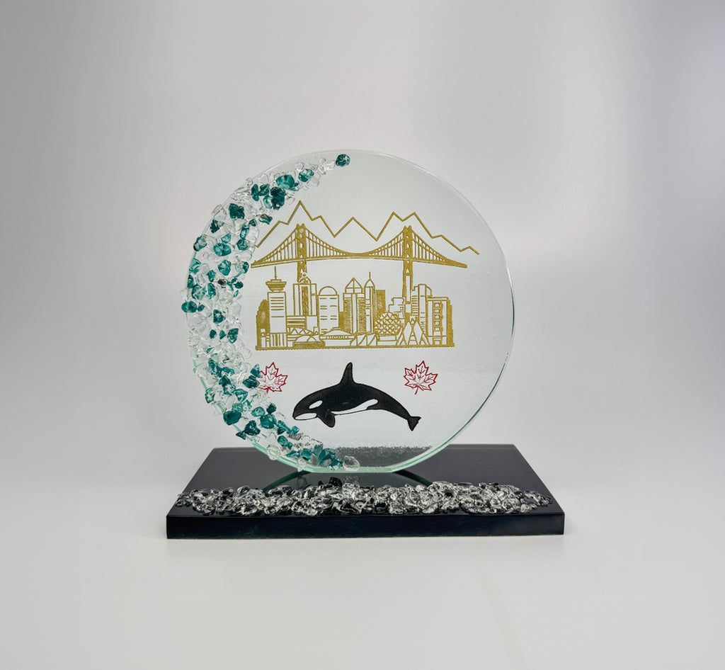 6 inch tall handcrafted glass award with colored glass gems fused into the front of the award in the shape of a crescent moon. the award has an multi colored etching of Vancouver landmarks, Canadian maple leafs and an orca killer whale. the award has a black glass base with clear glass gems fused into the base. the award stands in front of a studio background