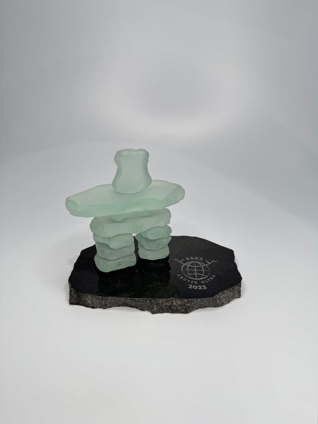 handcrafted 3.5 inch tall frosted glass inukshuk statue, on a chipped edge polished black granite base, with etching on the base. in front of a studio white background. 