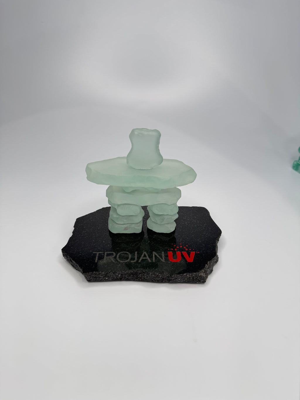 handcrafted 3.5 inch tall frosted glass inukshuk statue, on a chipped edge polished black granite base, with etching on the base. in front of a studio white background. 
