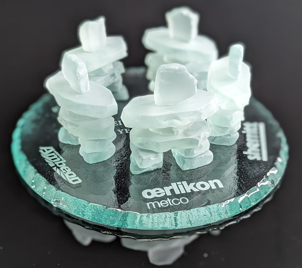 5 Frosted glass inukshuks standing in a friendship circle. on top of a glass base with logos engraved behind each inukshuk
