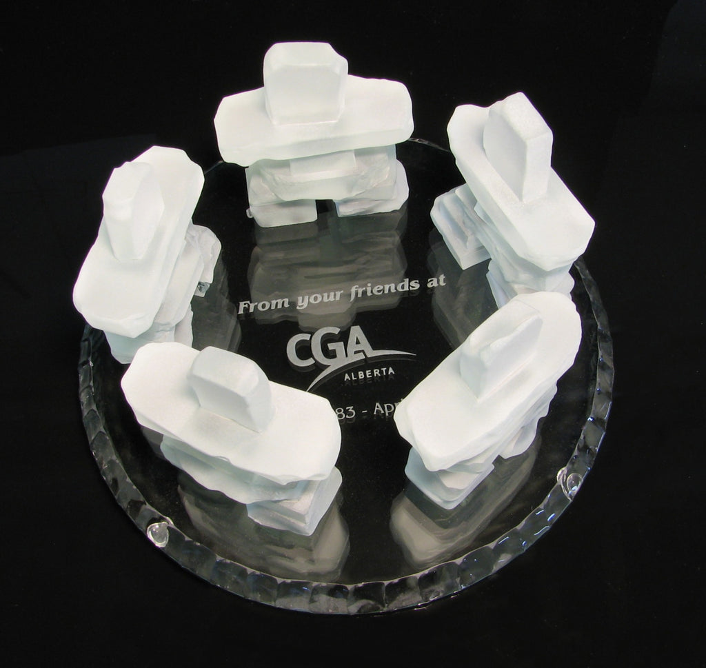 Five Frosted glass Inukshuks standing in a circle on top of a glass base with the engraving "from your firiends at cga alberta" on the base